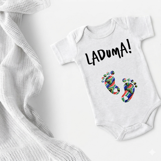 A white short-sleeve baby bodysuit laid flat on a white background, printed with the word 'LADUMA!' in black and two colorful baby footprints. A white textured knit blanket is draped along the left side.