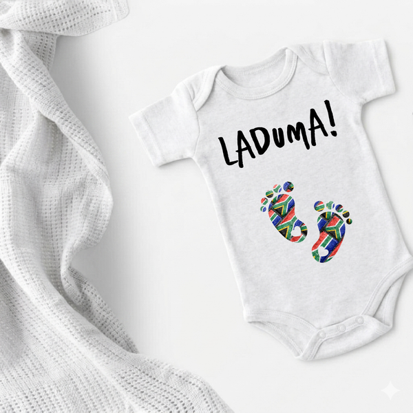 A white short-sleeve baby bodysuit laid flat on a white background, printed with the word 'LADUMA!' in black and two colorful baby footprints. A white textured knit blanket is draped along the left side.